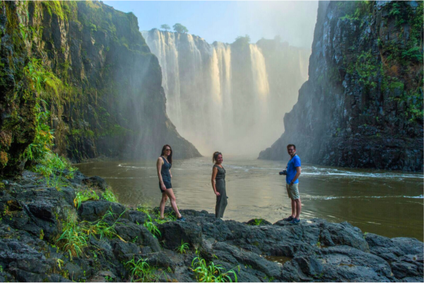 Three individuals stand on rocks directly in front of a majestic waterfall, with its powerful cascade of water.