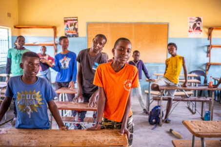 A group of boys standing in a classroom, some leaning on desks and others standing in a relaxed manner