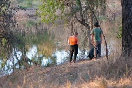 Two volunteers stand by a river, with a large tree in the background.
