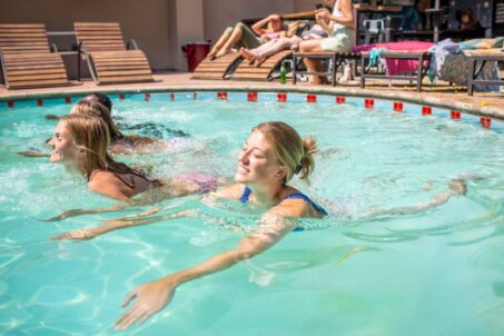 oung women enjoying a swim in a sunlit pool, with others relaxing poolside.